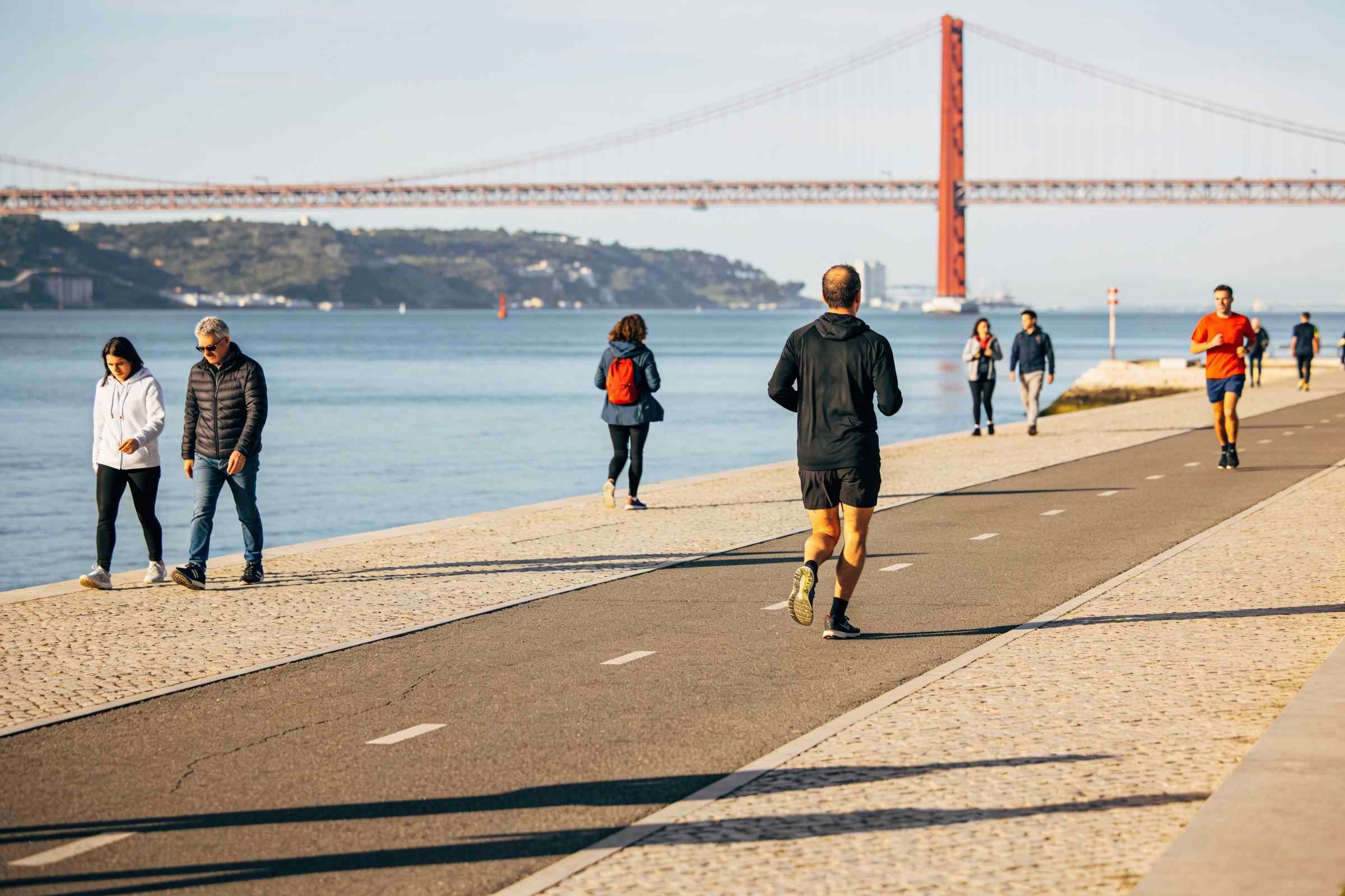 Runners on a riverside promenade with the water and a long suspension bridge in the distance